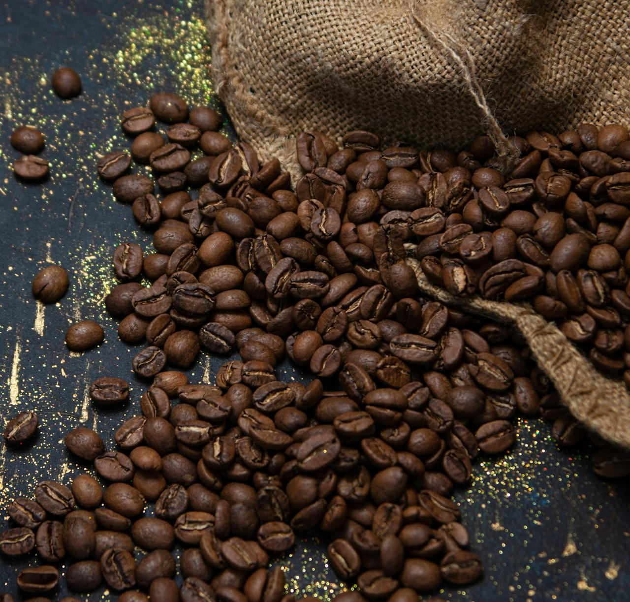 gallery-1 Heap of roasted coffee beans spilling from burlap sack on black background.