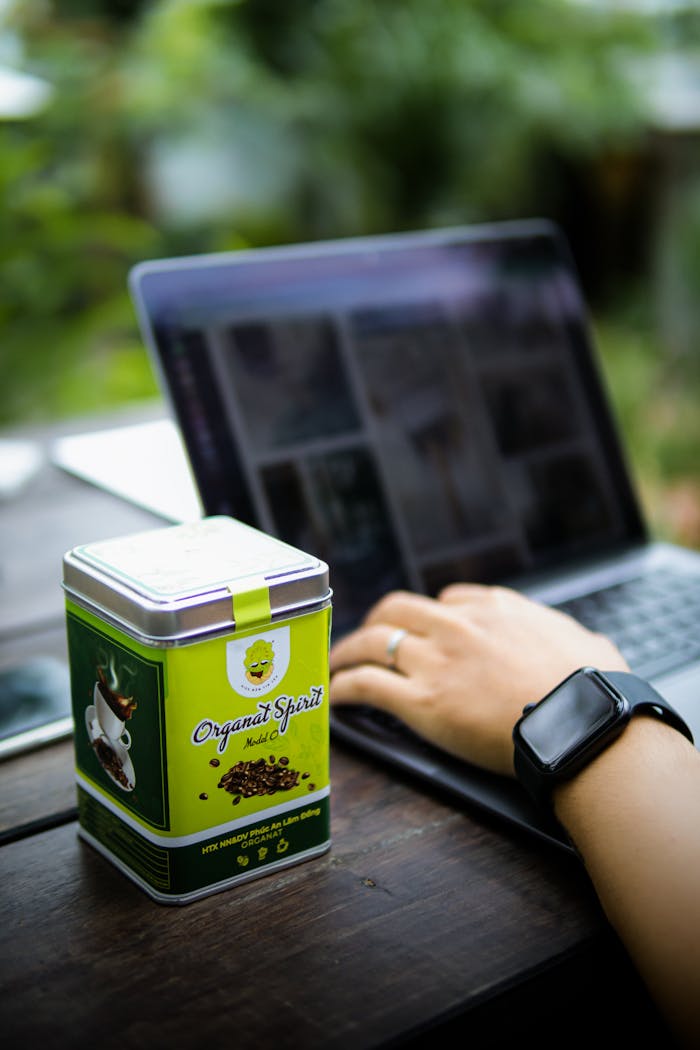 Close-up of a person working on a laptop next to a coffee tin outdoors.