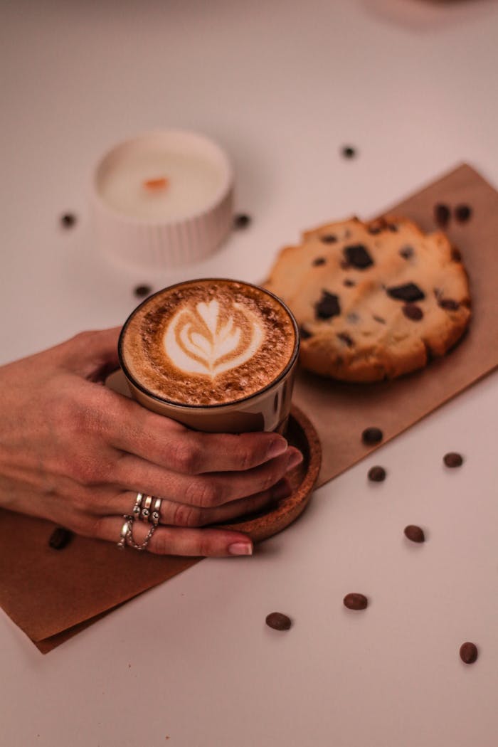 gallery-6 Aesthetic flat lay of cappuccino with latte art, chocolate chip cookie, and a candle on a wooden table.