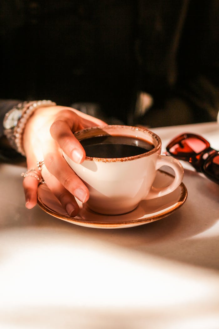 services-01 Close-up of a hand holding a coffee cup on a sunlit table with sunglasses and jewelry.