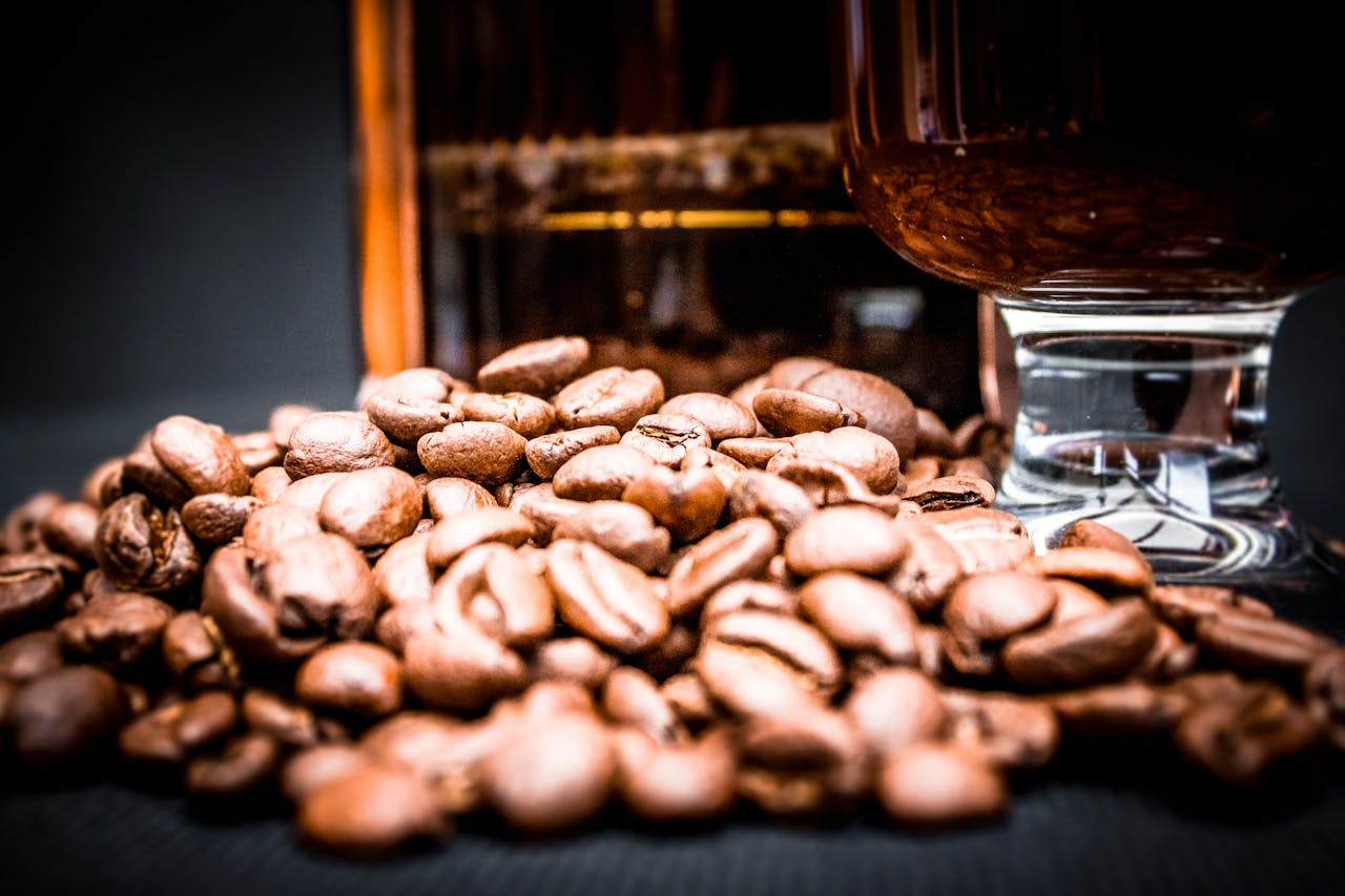 hero-img-02 Closeup of pile of aromatic coffee beans with glass of coffee and blurred French press behind on black background